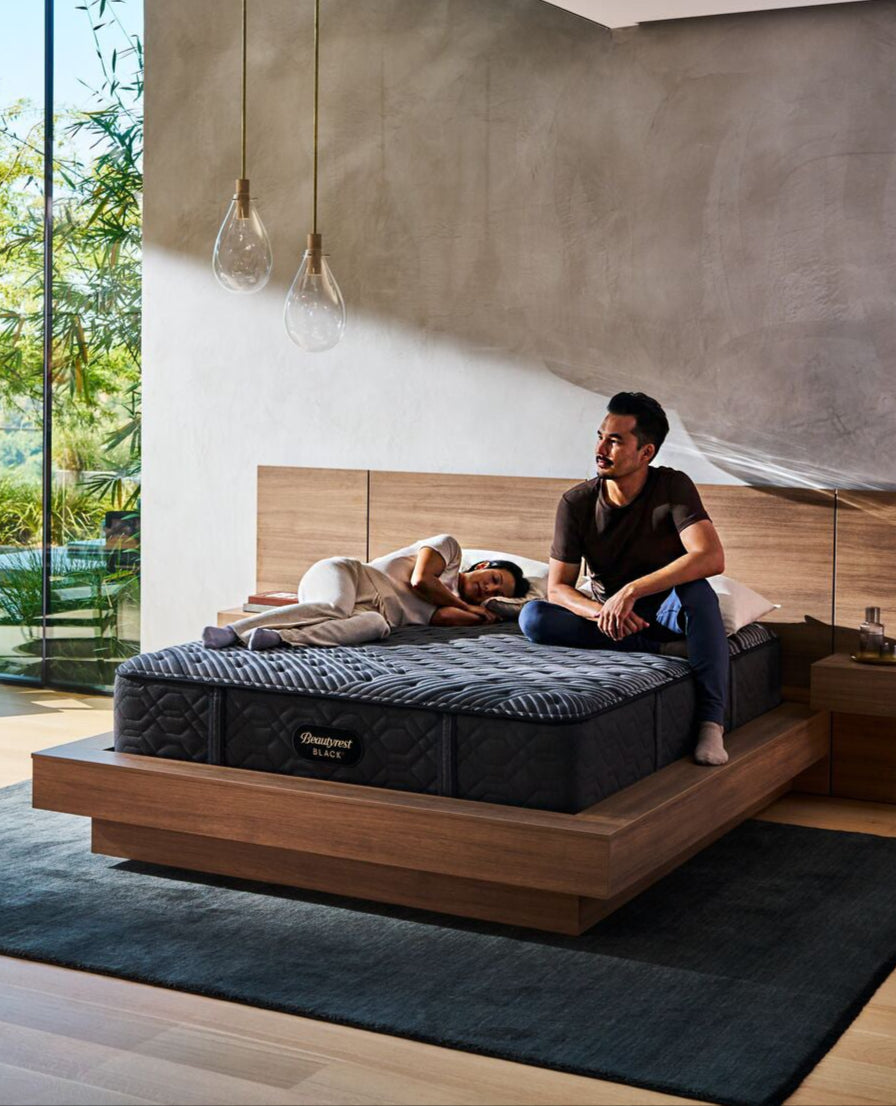 A man sitting on the edge of a Beautyrest Black mattress while a woman sleeps next to him, in a modern bedroom with a wooden bed frame, pendant lighting, and natural sunlight streaming in